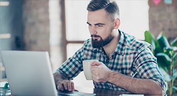 Man in checkered shirt holding a white mug looking at laptop